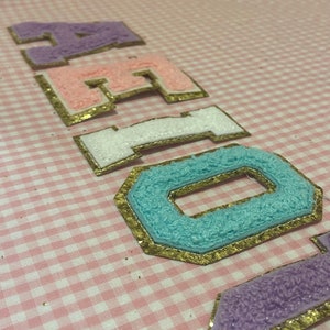 May include: A close-up of the word "HELLO" spelled out in colorful felt letters with gold glitter trim. The letters are arranged in a row on a pink and white gingham background.