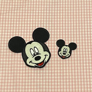 May include: Two black and white embroidered iron-on patches of Mickey Mouse. The larger patch is a close-up of Mickey's face with a big smile. The smaller patch is a smaller version of Mickey's face.