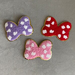 May include: Three colorful bow-shaped patches. One is purple with white polka dots, another is red with white polka dots, and the third is pink with white polka dots. Each bow has a gold glitter border.