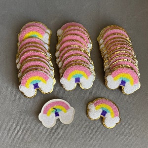 May include: A set of 10 rainbow-colored felt patches with a gold glitter outline. The patches are shaped like rainbows with a cloud at the bottom. The patches are perfect for adding a touch of whimsy to any project.