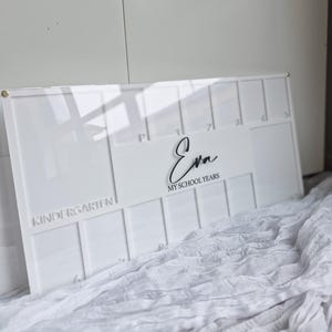 May include: A white acrylic school years keepsake board with the name "Eva" in a cursive font. The board is divided into sections for each school year, from Kindergarten to 7th grade. The text "MY SCHOOL YEARS" is also displayed.
