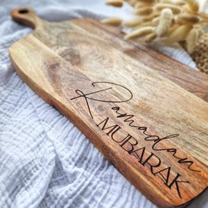 Könnte beinhalten: Ein Holzservierbrett mit Griff, mit dem eingravierten Text "Ramadan MUBARAK" in einer Schriftart. Das Brett hat eine natürliche Holzmaserung und eine hellbraune Farbe. Der Hintergrund ist ein weißes, strukturiertes Gewebe.