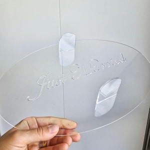 May include: A clear, oval acrylic sign with the engraved words "Just Married." The sign is held in place by two white adhesive pads. A wedding decoration.