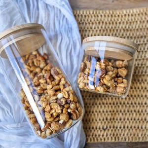 May include: Two glass jars with wooden lids filled with a mixture of nuts and candies. The jars are on a woven placemat and a white fabric.
