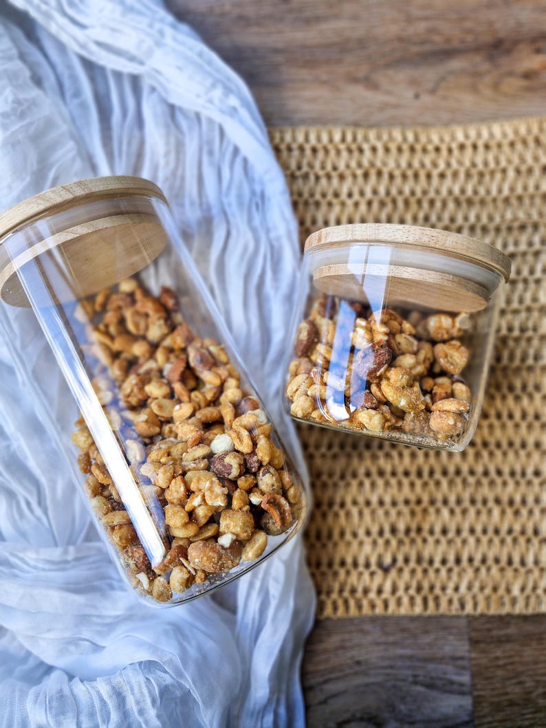 May include: Two glass jars with wooden lids filled with a mixture of nuts and candies. The jars are on a woven placemat and a white fabric.