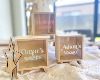 May include: Three wooden money boxes with clear glass fronts. Each box has a name on it: "Omar's Sadaqa Box", "Adam's Sadaqa Box", and "Mariam's Sadaqa Box". The boxes are on a white fabric surface with string lights.