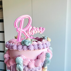 May include: A two-tiered pink cake decorated with pastel blue and purple frosting swirls. The cake is topped with a pink neon sign that says "Rena".
