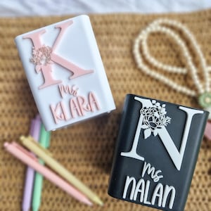 Può includere: Due custodie rettangolari personalizzate, una bianca con una "K" rosa e un design floreale, l'altra nera con una "N" bianca e un design floreale. Entrambe le custodie hanno nomi e il titolo "Ms." in corsivo. Penne pastello sullo sfondo.