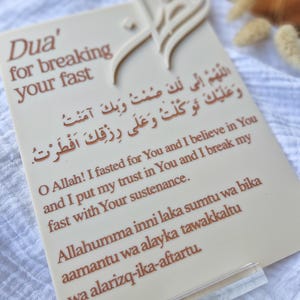 Può includere: Una carta beige con testo marrone, con scrittura araba e traduzione inglese. La carta recita "Dua' per rompere il tuo digiuno". Un ornamento decorativo è in alto. La carta è tenuta da un supporto in acrilico trasparente.