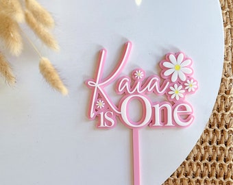 May include: Pink cake topper with the text "Kai is One" in white lettering and white flowers.