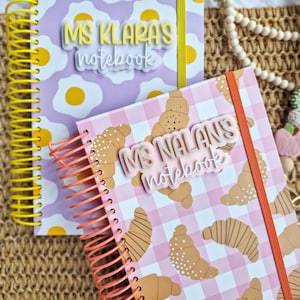 May include: Two spiral-bound notebooks. One has a purple background with egg graphics and a yellow spiral. The other features a pink gingham pattern with croissant graphics and an orange spiral binding. Each has custom text.