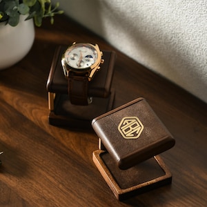 May include: A gold-toned watch with a brown leather band rests on a dark brown watch stand. A second watch stand with a gold emblem is also visible. The stands are made of wood and leather, showcasing a classic design.