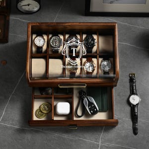 May include: A dark brown wooden watch box with a glass lid and a pull-out drawer. The top section features ten compartments with beige cushions for watches. The drawer contains smaller dividers holding coins, a white case, and a car key fob.