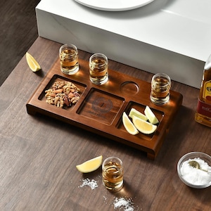 May include: A wooden serving tray with four shot glasses, a central engraved area, and compartments for nuts and lemon wedges. The tray is on a wooden surface, with a bottle of tequila and a small bowl of salt.