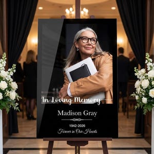 May include: A black memorial sign featuring a photograph of a person with gray hair and glasses holding a laptop. The text reads In Loving Memory, Madison Gray, 1942-2026, and Forever in Our Hearts, displayed on a wooden easel stand.