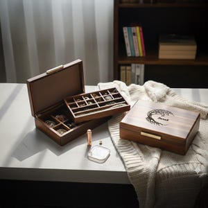 May include: Wooden jewelry box with a hinged lid and a gold-colored clasp. The box is open, revealing compartments for jewelry. A smaller tray with jewelry sits inside. A closed box with a tree design and the name "Sarah" is also present.