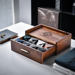 May include: A dark wood storage box with a pull-out drawer containing multiple folded neckties. The top of the box features a laser-engraved crest with the text Thomas Anderson. The drawer has a metallic handle and holds ties in various patterns.