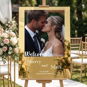 May include: A gold-framed wedding sign on an easel. The sign features a photo of a kissing couple, with the words "Welcome to our Wedding" and the names "Sherry and Mark" along with the date and location.