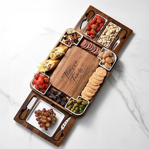 Puede incluir: Una tabla de embutidos rectangular de madera con una sección central grabada con el texto Thompson Family. La tabla incluye varios platos de servicio de cerámica blanca llenos de fresas, nueces, uvas, aceitunas, salami y galletas.