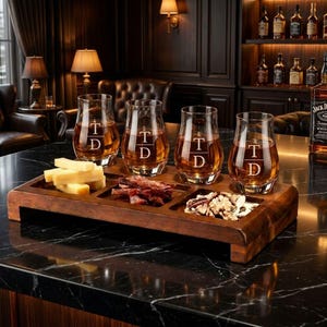 A wooden tray displaying four glasses filled with whiskey, accompanied by various snacks, including cheese and crackers, on a marble countertop.