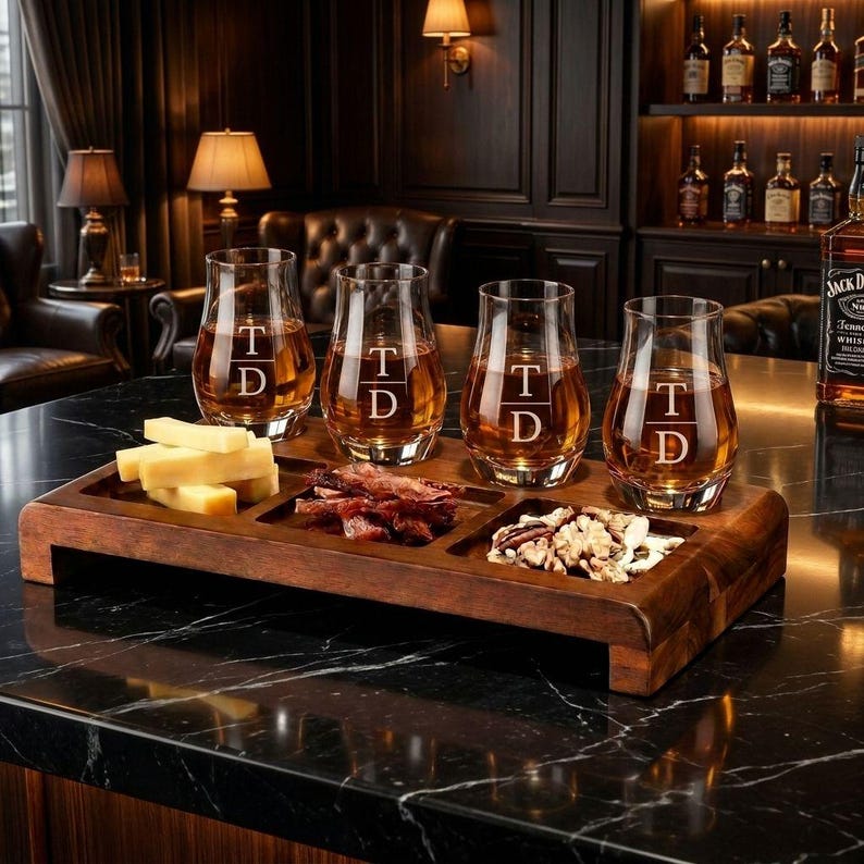 A wooden tray displaying four glasses filled with whiskey, accompanied by various snacks, including cheese and crackers, on a marble countertop.