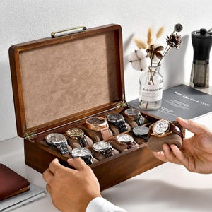 May include: A wooden watch box with a hinged lid, holding several wristwatches with various band and face designs. The box is open, revealing a tan interior and a brass handle. A hand is holding a watch.