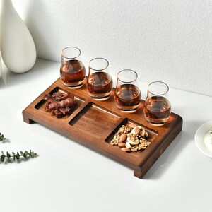 A wooden tray with four small glasses containing different types of drinks, accompanied by a small plate of nuts, is displayed on a white surface.