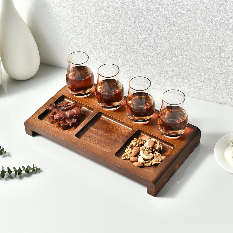 A wooden tray with four small glasses containing different types of drinks, accompanied by a small plate of nuts, is displayed on a white surface.
