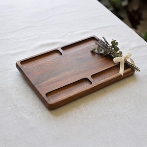 Puede incluir: Una bandeja rectangular de madera con acabado marrón oscuro. La bandeja tiene tres secciones empotradas y un ramo decorativo de lavanda seca y vegetación atado con una cinta crema. La bandeja está sobre una superficie blanca.