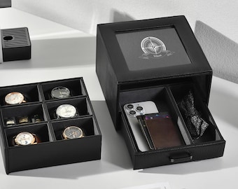 Könnte beinhalten: Eine schwarze, strukturierte Uhrenaufbewahrungsbox mit Glasdeckel, der eine weiße Kompassgrafik und den Namen Wilson zeigt. Die Box enthält ein herausnehmbares Fach mit sechs Uhrenplätzen sowie eine Schublade mit Smartphone, Geldbörse und Fliege.
