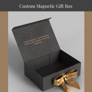 May include: A black custom magnetic gift box with a gold ribbon and bow. The box is open, revealing a message that reads, "Congratulations on your wedding! Wishing you a lifetime of love and happiness."
