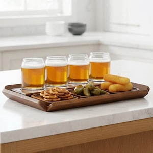 May include: A rectangular wooden serving tray featuring four glass beer cans filled with amber liquid and white foam, alongside three small square wooden bowls containing pretzels, green olives, and golden fried snacks.