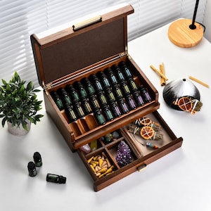 Könnte beinhalten: Eine Aufbewahrungsbox aus dunklem Holz mit Klappdeckel und Schublade. Der obere Bereich enthält vierundzwanzig kleine Fläschchen mit ätherischen Ölen aus dunklem Glas. Die untere Schublade ist mit Räucherkegeln, einem violetten Kristall und Steinen gefüllt.
