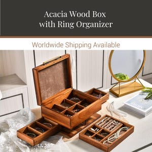 a wooden jewelry box with multiple compartments, showcasing various pieces of jewelry. The box is placed on a table, and there is a mirror and a book nearby. The image also includes text that reads "Acacia Wood Box with Ring Organizer" and "Worldwide Shipping Available".