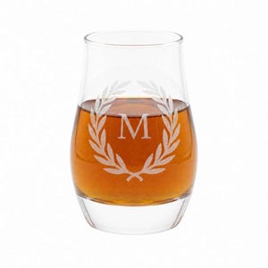 A glass of amber-colored liquid, possibly whiskey, with a white laurel wreath engraved on it.