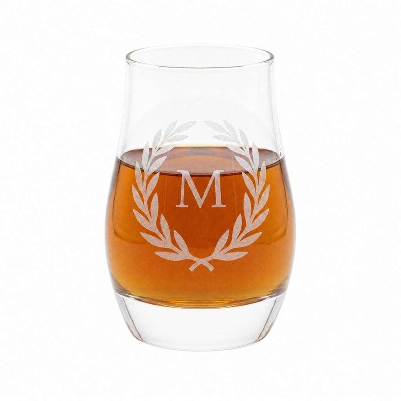A glass of amber-colored liquid, possibly whiskey, with a white laurel wreath engraved on it.