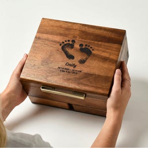 Puede incluir: Caja de recuerdos de madera oscura con huellas de bebé y el nombre "Emily" con fecha y hora. La caja tiene un asa dorada y mide 3,3 kg / 58,4 cm.