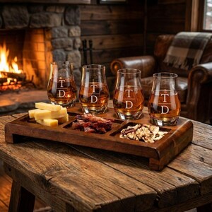 A wooden tray with four glasses filled with amber-colored liquid, likely whiskey, and various snacks such as cheese, nuts, and dried fruit, placed on a wooden table in front of a fireplace.