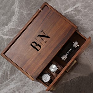 May include: A rectangular dark brown wooden storage box with a hinged lid featuring the black engraved letters B|N. The open drawer reveals two silver-toned wristwatches with white faces and a black folding pocket knife with silver metal accents.