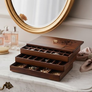 Peut inclure: Une bo&icirc;te &agrave; bijoux en bois &agrave; trois niveaux avec une finition marron fonc&eacute;. Le couvercle sup&eacute;rieur pr&eacute;sente une gravure florale et un petit fermoir dor&eacute;. Les tiroirs ouverts r&eacute;v&egrave;lent des compartiments pour ranger les bijoux. Un miroir dor&eacute; est en arri&egrave;re-plan.