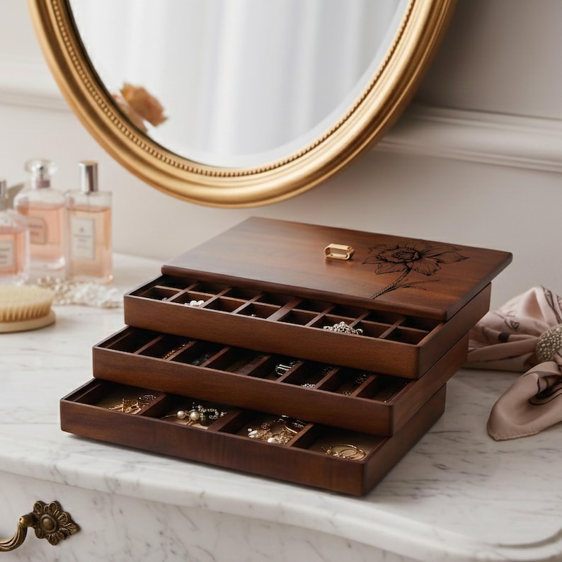 Peut inclure: Une bo&icirc;te &agrave; bijoux en bois &agrave; trois niveaux avec une finition marron fonc&eacute;. Le couvercle sup&eacute;rieur pr&eacute;sente une gravure florale et un petit fermoir dor&eacute;. Les tiroirs ouverts r&eacute;v&egrave;lent des compartiments pour ranger les bijoux. Un miroir dor&eacute; est en arri&egrave;re-plan.