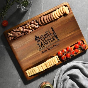 Puede incluir: Tabla de servir rectangular de madera con tres secciones empotradas. La tabla está grabada con "The Grill Master" y "Your Text Here". Contiene galletas saladas, galletas, anacardos y tomates cherry. Sobre una superficie gris.