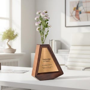 Puede incluir: Un jarrón de madera con diseño geométrico, con una placa de bronce que muestra "Leadership & Vision" y un nombre, sosteniendo un ramo de flores blancas y moradas. El jarrón está sobre un escritorio blanco.