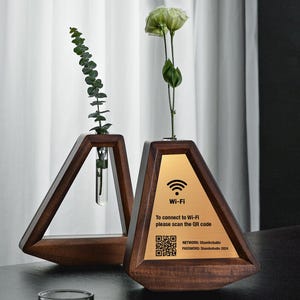 May include: Two wooden triangular vases with a dark finish. One holds a sprig of greenery in a glass tube, the other has a gold plate with Wi-Fi instructions and a QR code. The vases are on a dark surface.