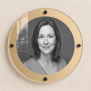 May include: A circular wall display featuring a black and white portrait of a person with shoulder-length hair. The portrait is encased between a clear acrylic front panel and a gold-toned circular back plate, secured by four black metal standoffs.