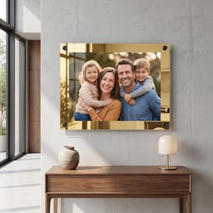 May include: A rectangular wall-mounted photo frame featuring a reflective gold border and four black corner mounting studs. The frame displays a color photograph of a family of four, including two adults and two children, smiling together.