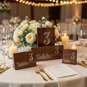 May include: Three dark wood table number signs with white engraved text. The signs feature the number 3 and the word table in a script font. They are arranged on a white tablecloth alongside a floral centerpiece, candles, and place settings.