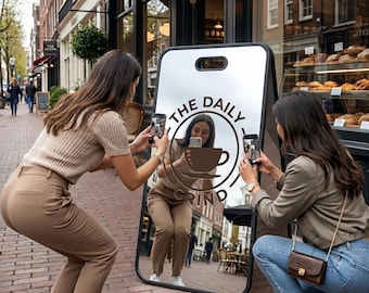 Puede incluir: Espejo en forma de caballete con el texto "THE DAILY GRIND" y un logotipo de taza de café. Dos personas están tomando fotos del espejo con sus teléfonos. El espejo refleja una escena callejera con una panadería.