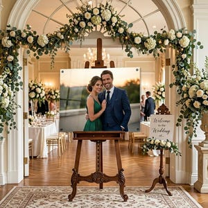 Puede incluir: Un caballete de madera exhibe una foto enmarcada de una pareja en una boda. La foto est&aacute; colocada contra un fondo de un arco decorado con flores blancas y vegetaci&oacute;n. Tambi&eacute;n hay un letrero que dice "Bienvenida a la boda de..."