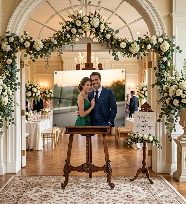 Puede incluir: Un caballete de madera exhibe una foto enmarcada de una pareja en una boda. La foto est&aacute; colocada contra un fondo de un arco decorado con flores blancas y vegetaci&oacute;n. Tambi&eacute;n hay un letrero que dice "Bienvenida a la boda de..."